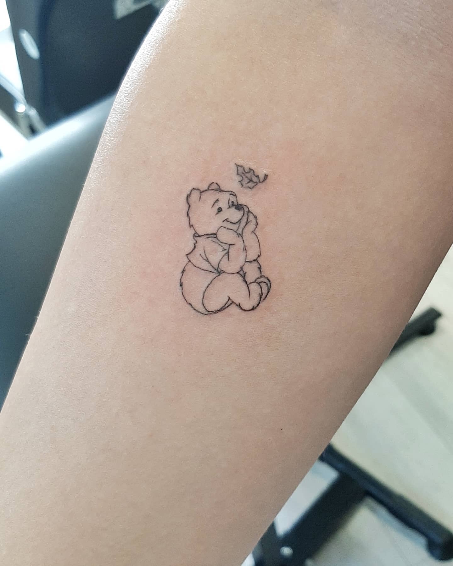 Small tattoo of Winnie the Pooh sitting and gazing at a falling leaf on a person's forearm